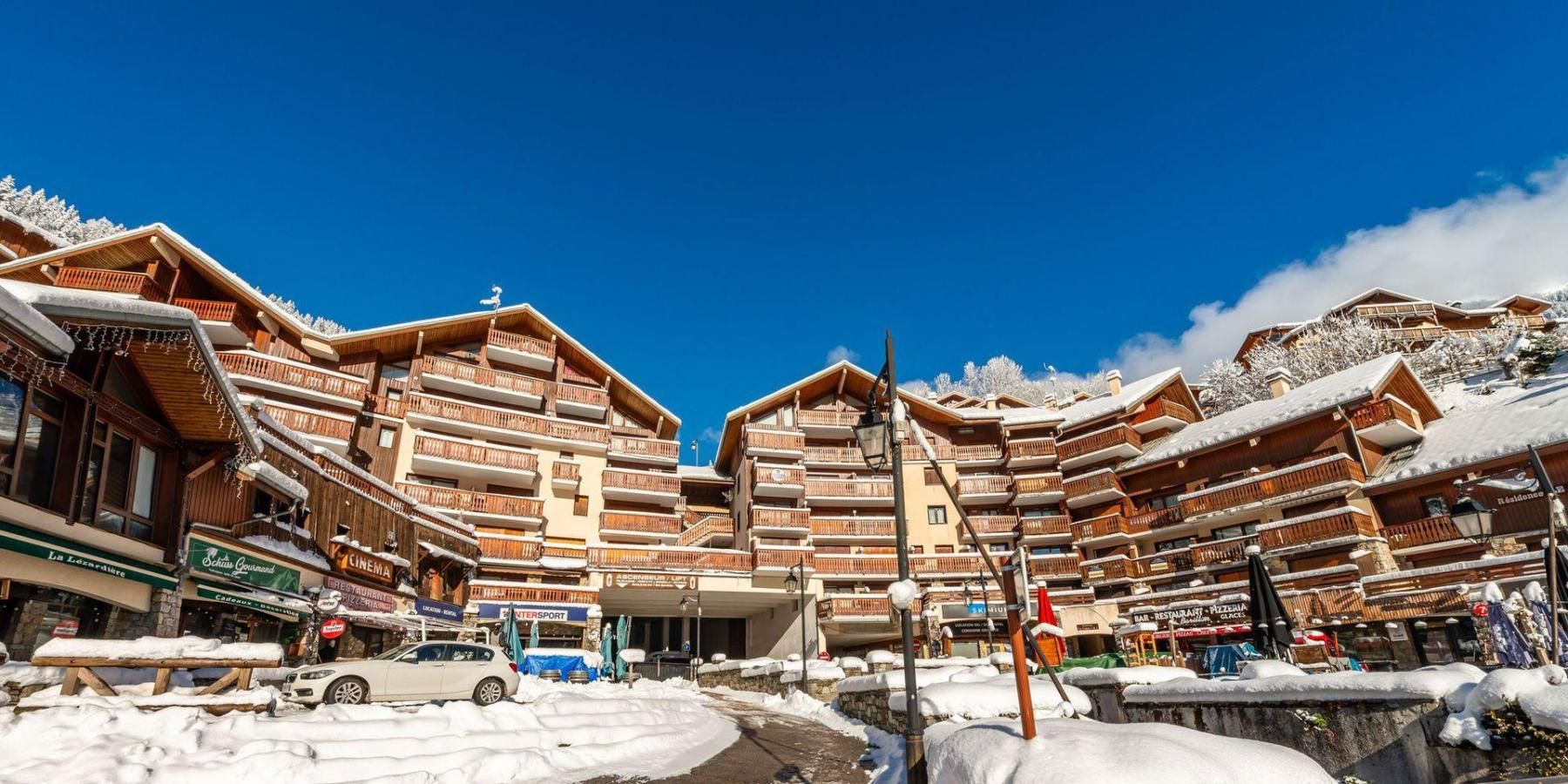 A snow-covered mountain resort with multi-story wooden buildings, a clear blue sky, and a paved street.