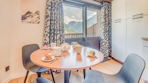 A dining area with a round table, chairs, and a view of mountains through a window with floral curtains. - Resort center, 4th floor, balcony, tv, 29m², Champagny-en-Vanoise (Champagny-en-Vanoise)