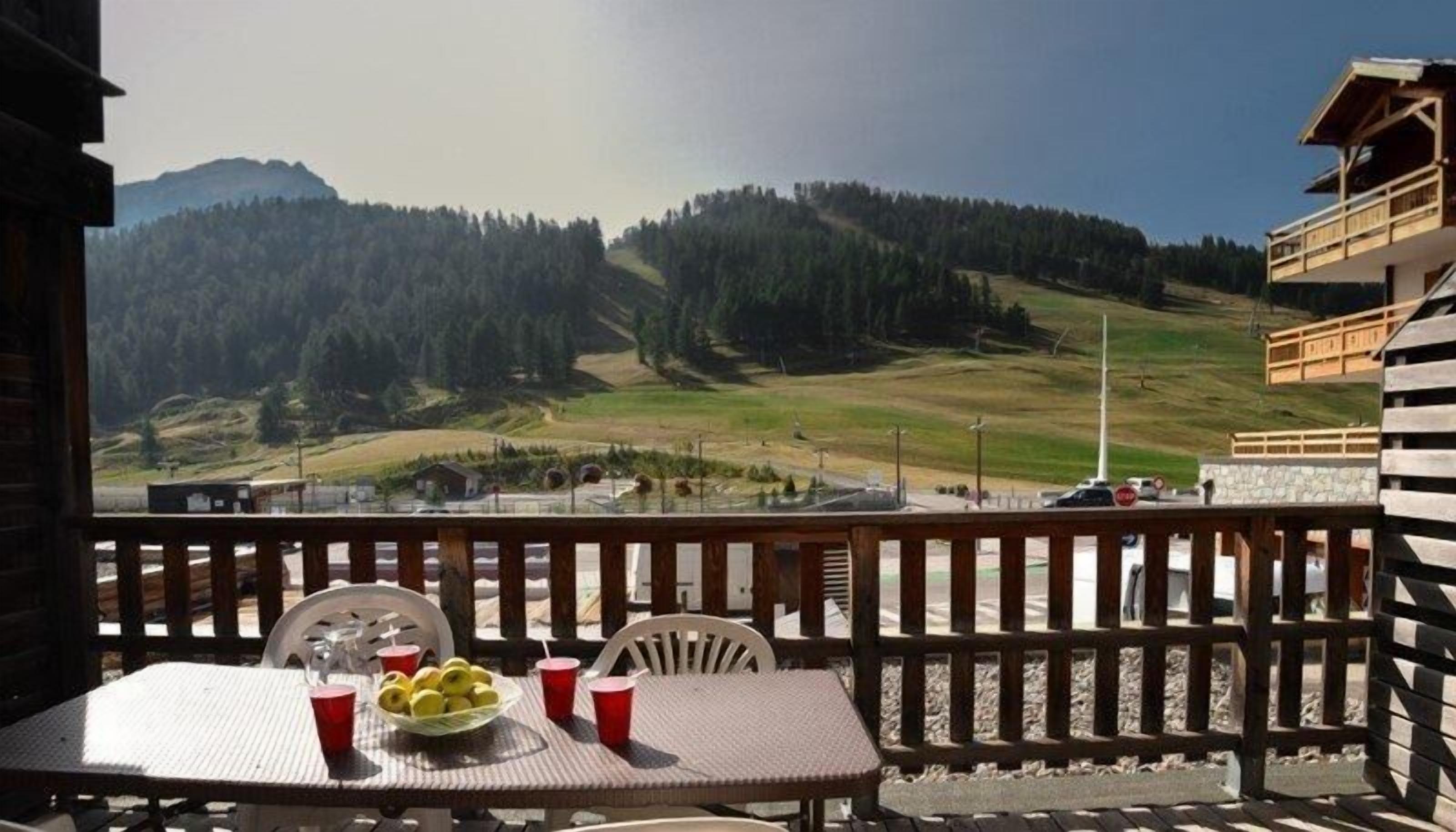 A balcony with a table set for two, a basket of fruit, and red cups. A mountain landscape is visible in the background.