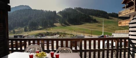 A balcony with a table set for two, a basket of fruit, and red cups. A mountain landscape is visible in the background.