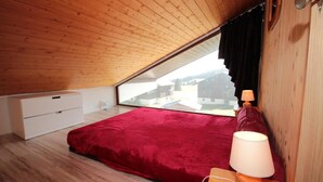 A bedroom with a sloped ceiling, a bed with a red bedspread, a bedside table with a lamp, and a window overlooking a village. - 300m from ski slopes, 2nd floor, view panoramic, balcony, tv, ski locker, 45m², Les Saisies (Les Saisies)