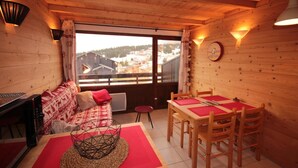 A cozy room with a wooden interior, a sofa with patterned cushions, a dining table with chairs, and a view of a village through a large window. - 300m from ski slopes, 2nd floor, view panoramic, balcony, tv, ski locker, 45m², Les Saisies (Les Saisies)