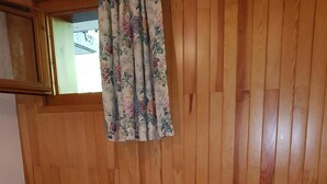 Ein Zimmer mit Holzwandverkleidung, einem Bett, zwei Nachttischlampen und einem Fenster mit blumigen Vorhängen.