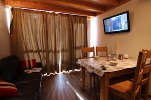Um quarto aconchegante com teto de madeira, uma mesa de jantar com cadeiras, uma televisão na parede e uma janela com cortinas.