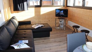 Uma sala de estar moderna com um sofá de couro preto, uma mesa de centro de vidro, uma TV de tela plana e um pequeno espaço de cozinha com pia e banquetas.