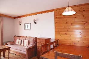 A room with a wooden wall, a sofa with patterned cushions, a wooden dining table with chairs, and a hanging light fixture.