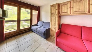 A room with a large sliding glass door leading to a grassy area, a grey sofa, a red sofa, and wooden cabinets. - 10m from ski slopes, 10th floor, view ski slopes, swimming pool, balcony, tv, ski locker, 22m² (Le Corbier)