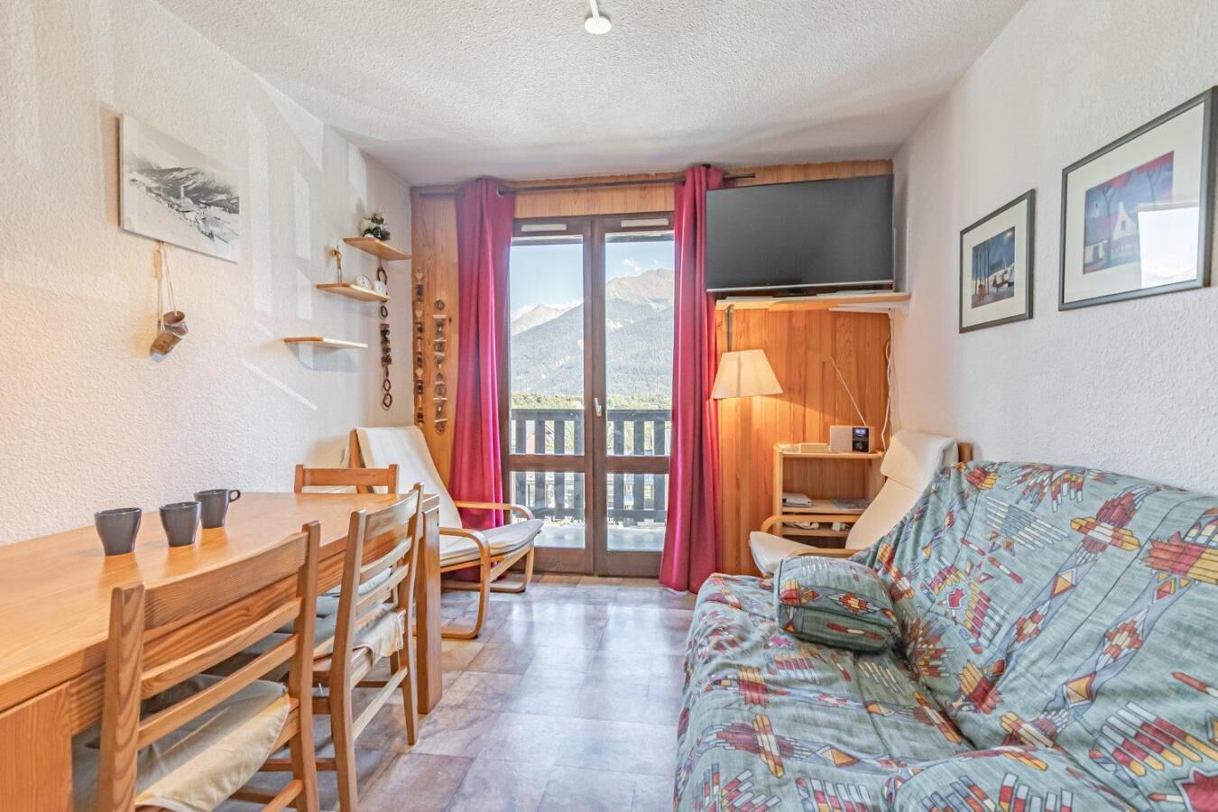 Une pièce chaleureuse avec une table à manger en bois, des chaises, un canapé à motifs, un téléviseur et une vue sur les montagnes, visible à travers une porte vitrée.