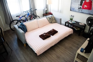 Ein ordentlich eingerichtetes Schlafzimmer mit karierter Bettdecke, einem grauen Sofa, einem Fahrrad und einem kleinen Tisch mit Lampe und Dekoration.