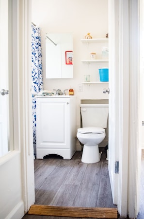 Ein kleines, schmales Badezimmer mit einer weißen Toilette, einem weißen Waschbecken und einem Spiegel darüber. Über der Toilette befinden sich Regale mit verschiedenen Gegenständen.
