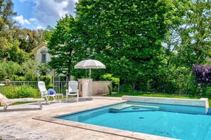Une piscine dotée de chaises longues et d’un parasol, entourée de verdure et d’un bâtiment.