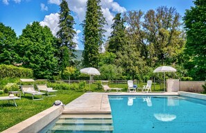 Piscina con lettini e ombrelloni, circondata da vegetazione e alberi.