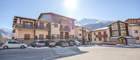 A multi-story building with balconies, a parking lot, and snow-covered mountains in the background.