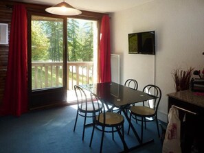 A dining area with a table and chairs, a television on the wall, and a view of trees through a sliding door.