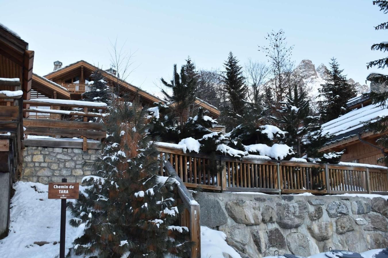 A wooden chalet with stone walls, snow-covered roofs, and a sign indicating a 'Chemin de TARA' pathway.