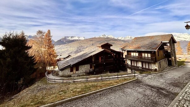 Un chalet de montagne à la façade en pierre et aux balcons en bois, situé au pied de sommets enneigés.