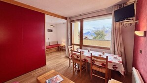 Une pièce chaleureuse avec une table à manger en bois, des chaises, un paravent rouge, une fenêtre donnant sur un beau paysage et un téléviseur fixé au mur.