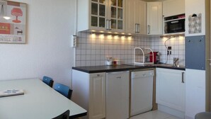 Eine kleine Küche mit weißen Schränken, einer Mikrowelle und einem Spülbecken. Ein Esstisch mit Stühlen und ein gerahmtes Bild an der Wand sind ebenfalls vorhanden.