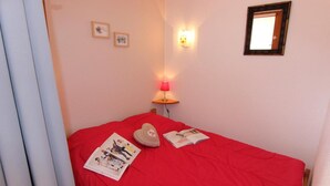Une petite pièce accueillante, avec une couvre-lit rouge, un coussin en forme de cœur et un magazine sur le lit. Il y a une lampe fixée au mur et un tableau encadré accroché au mur.