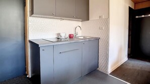 Een kleine keuken met grijze kastjes, een roestvrijstalen aanrecht en een rode waterkoker.