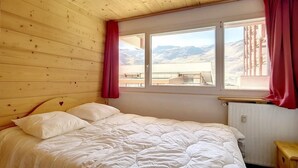 Een slaapkamer met een houten plafond, een bed met wit beddengoed, een raam met uitzicht op bergen en een radiator.