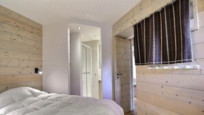 A bedroom with a wooden slat wall, a bed with white bedding, a window with striped curtains, and a door leading to another room. - 10m from ski slopes, 1st floor, view ski slopes, balcony, 40m², Méribel-Mottaret (Méribel - Les Allues)