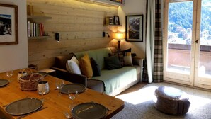 A cozy room with a wooden wall, a sofa with cushions, a dining table set for two, and a view of a snowy landscape through a window. - 10m from ski slopes, 1st floor, view ski slopes, balcony, 40m², Méribel-Mottaret (Méribel - Les Allues)