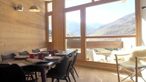 Uma sala de jantar com mesa e cadeiras de madeira, janelas amplas com vista para as montanhas e iluminação pendente.