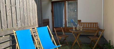 A small outdoor seating area with wooden furniture, including a bench and two chairs, and a table with a few glasses and a bottle.