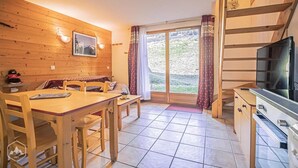 L’intérieur d’un chalet accueillant, avec une table à manger en bois, des chaises, un canapé, un téléviseur et une grande fenêtre agrémentée de rideaux.
