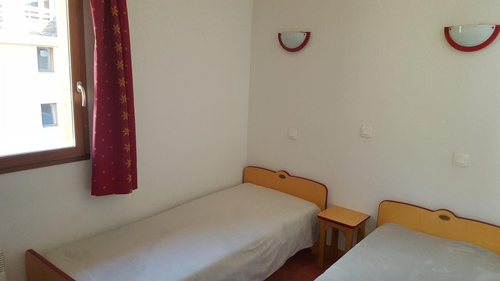 A small, simple room with two single beds, a small table, and a window with curtains.