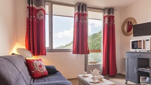 Ein gemütliches Zimmer mit einer Couch, einem kleinen Tisch mit Teekanne und Tassen, einem Fernseher auf einem Schrank und roten Vorhängen mit Blick auf die Berge.