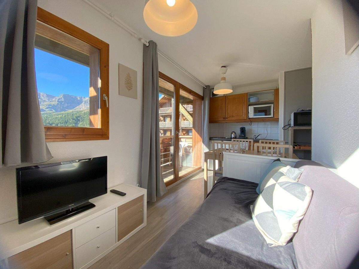 A compact room with a TV, a bed, a kitchenette, and a sliding glass door leading to a balcony.