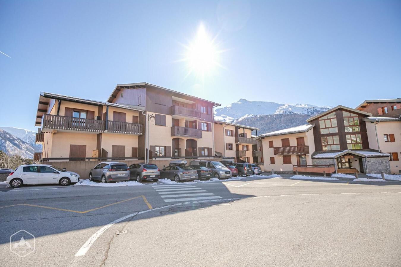 A multi-story building with balconies, a parking lot, and snow-covered mountains in the background.