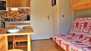 A compact kitchen with wooden cabinets, a microwave, and a coffee maker. There is a small dining table with a bowl and glasses. A door leads to a bedroom with a patterned bedspread.