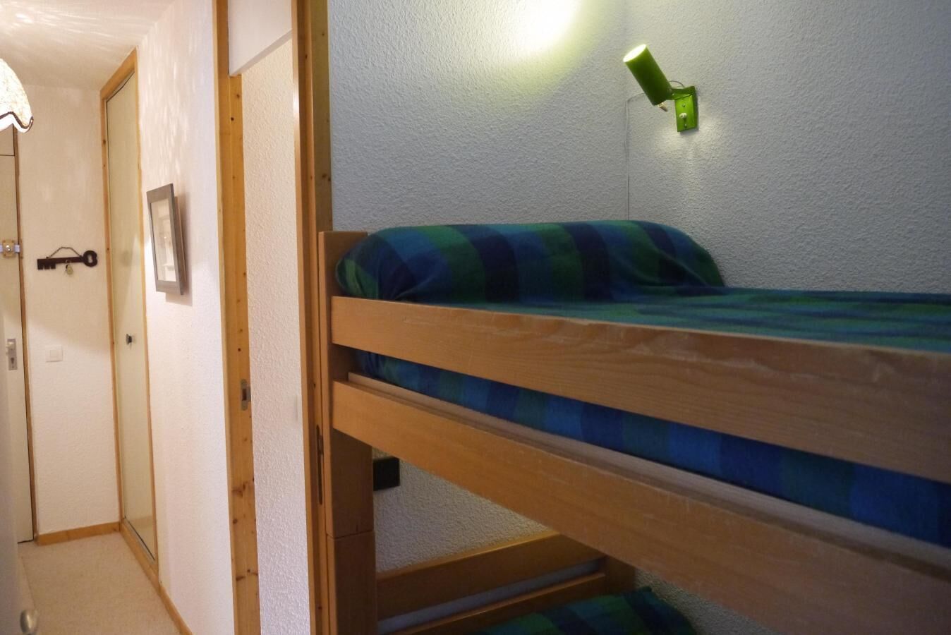 Una cama litera con una lámpara de pared verde, una puerta con cerradura y una pequeña foto enmarcada en la pared.
