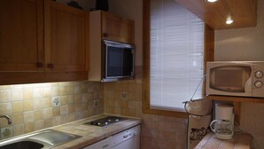 Une cuisine compacte avec des meubles en bois, un four à micro-ondes encastré, une machine à café et une fenêtre équipée de stores.