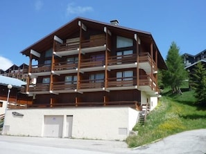 A multi-story wooden building with balconies, a garage, and a grassy slope. - 150m from resort center, 2nd floor, view ski slopes, balcony, tv, 35m², Les Saisies (Les Saisies)