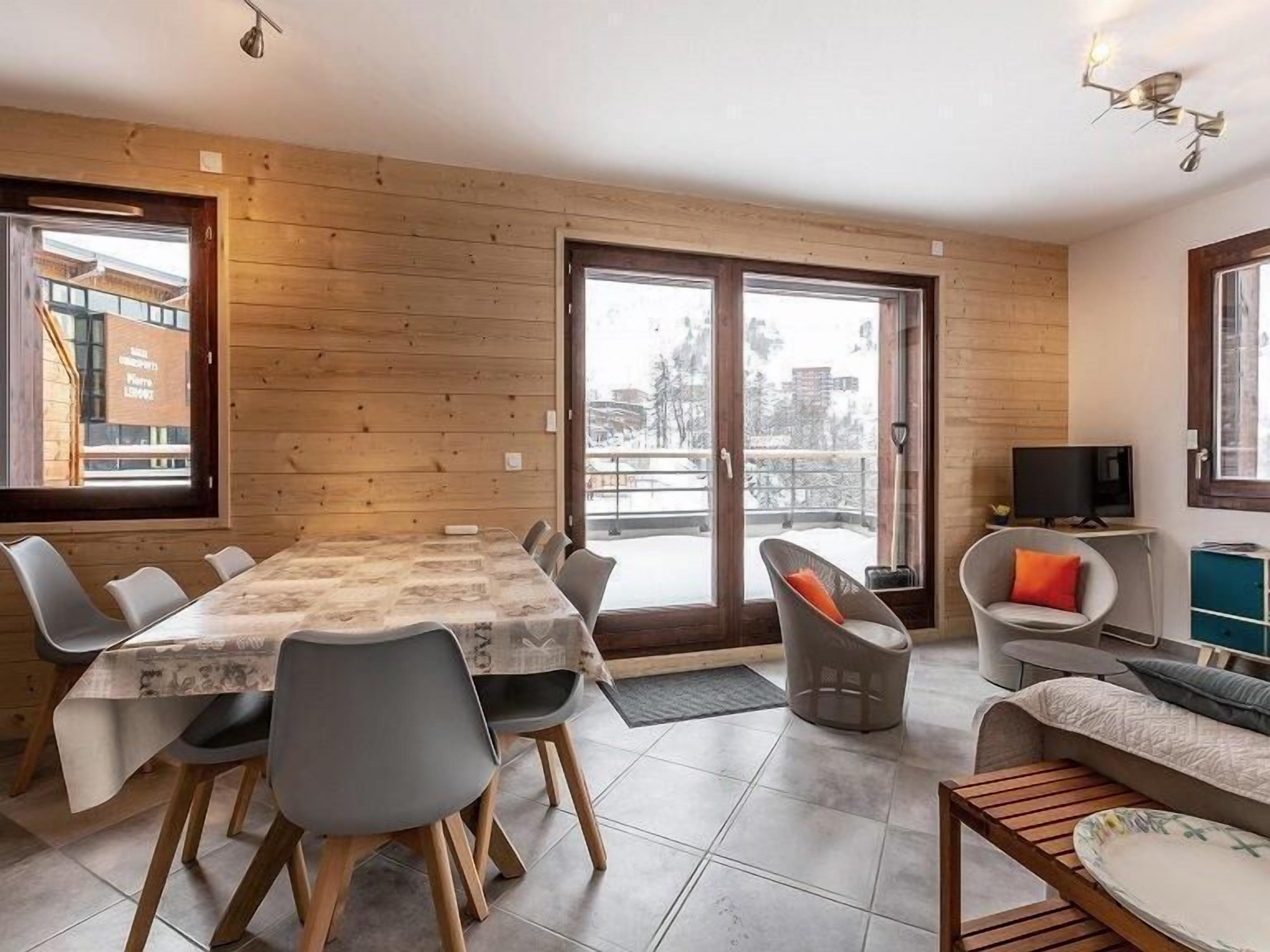 Ett modernt vardagsrum med träinredning, en matplats med bord och stolar, en soffa, en tv och utsikt över ett snötäckt landskap genom stora fönster.