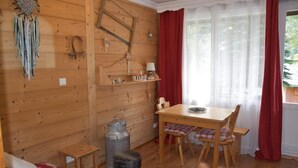 A cozy cabin interior with a wooden dining table, chairs, a small stool, a metal container, and a red and white patterned sofa.