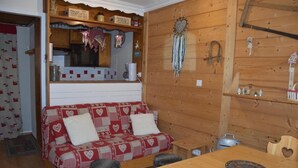 A cozy cabin interior with a wooden interior, a sofa with heart-patterned cushions, a small wooden table, and a kitchen area with a stove and cabinets.