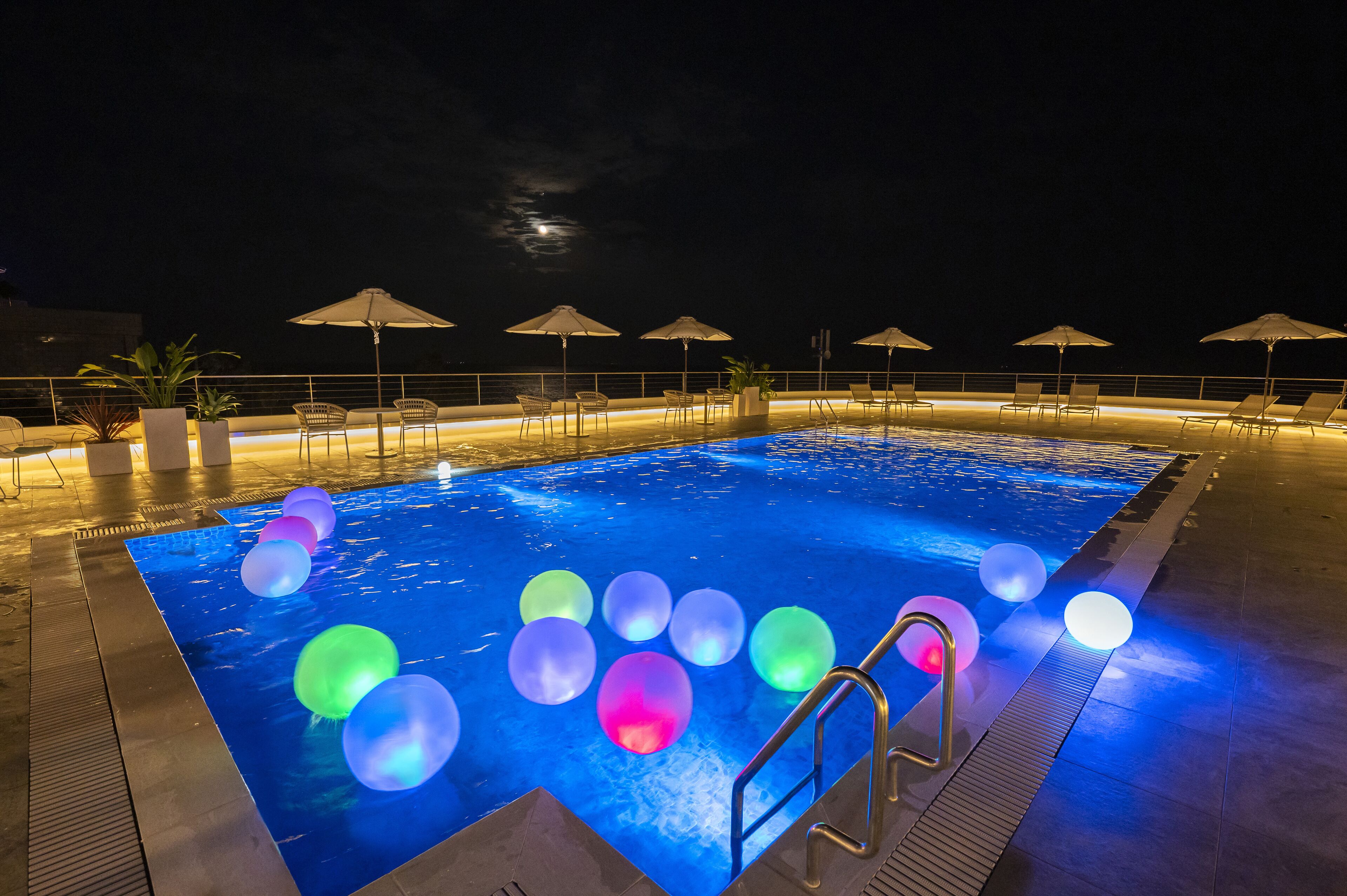 A pool area with a night sky view, illuminated water, and lounge chairs with umbrellas.