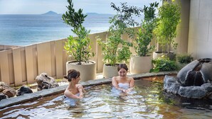 A hot spring area with two people in the water, surrounded by potted plants and a view of the sea. - Atagawa Ocean Resort (Higashiizu)