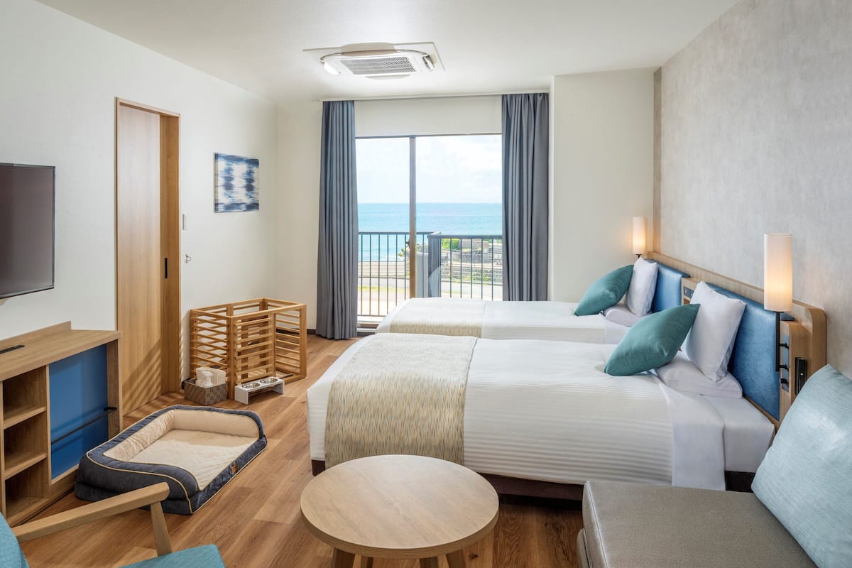 Habitación de hotel con dos camas, televisión, escritorio, silla, mesita y balcón con vistas al mar.