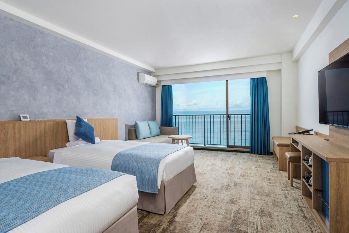 Habitación de hotel con dos camas, televisión, balcón con vistas y escritorio.