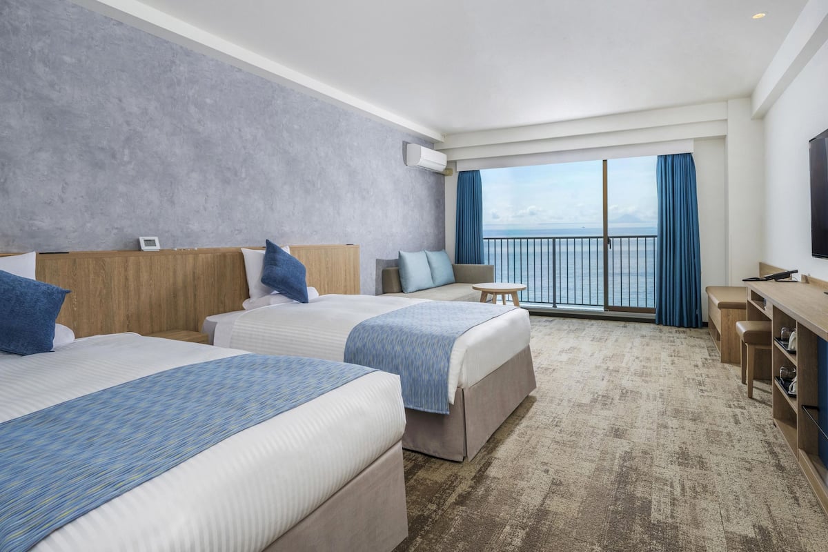 Habitación de hotel con dos camas, una mesita, televisor y vista al mar.