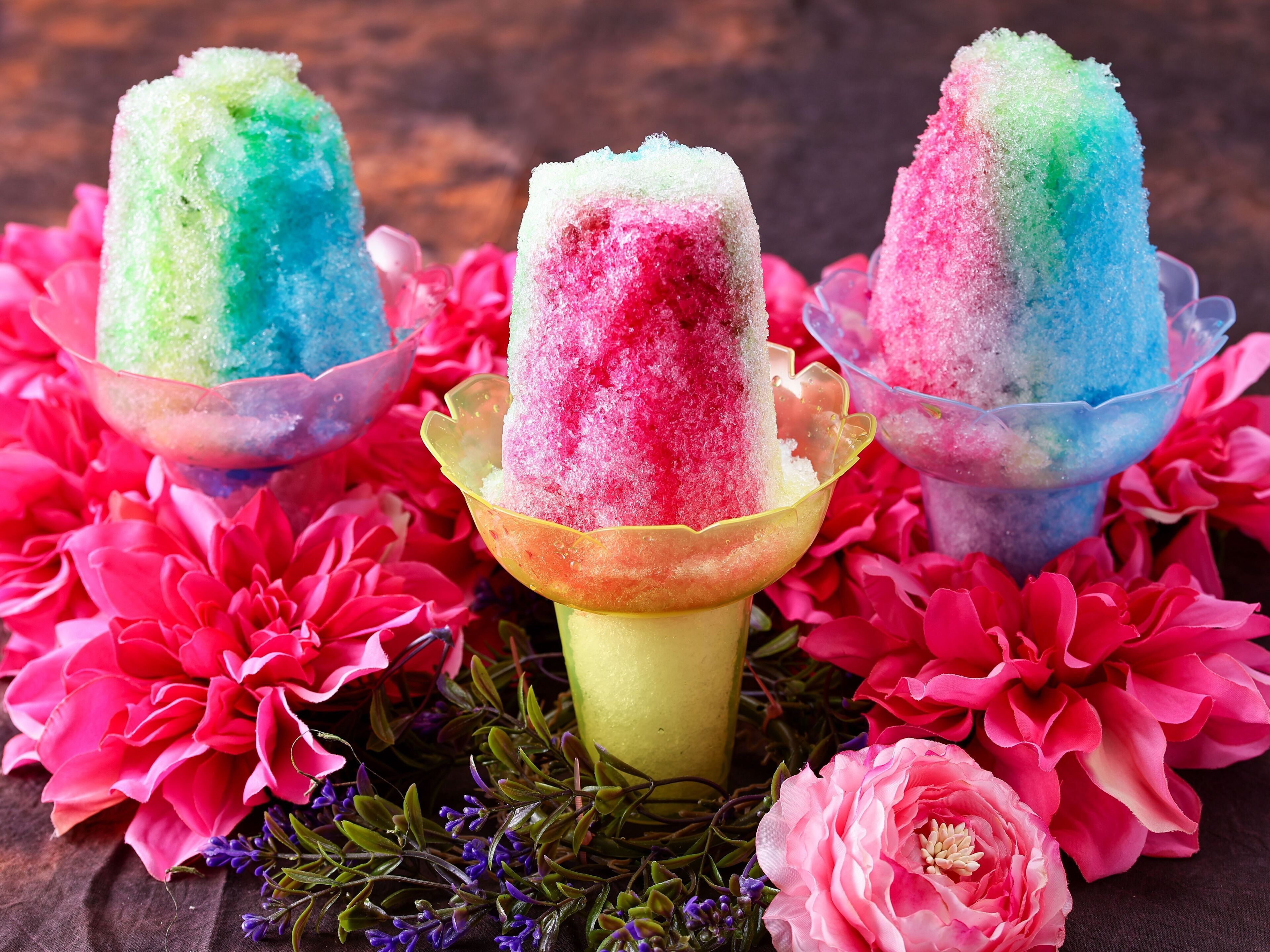 Three colorful popsicles in clear cups on a table surrounded by pink flowers.