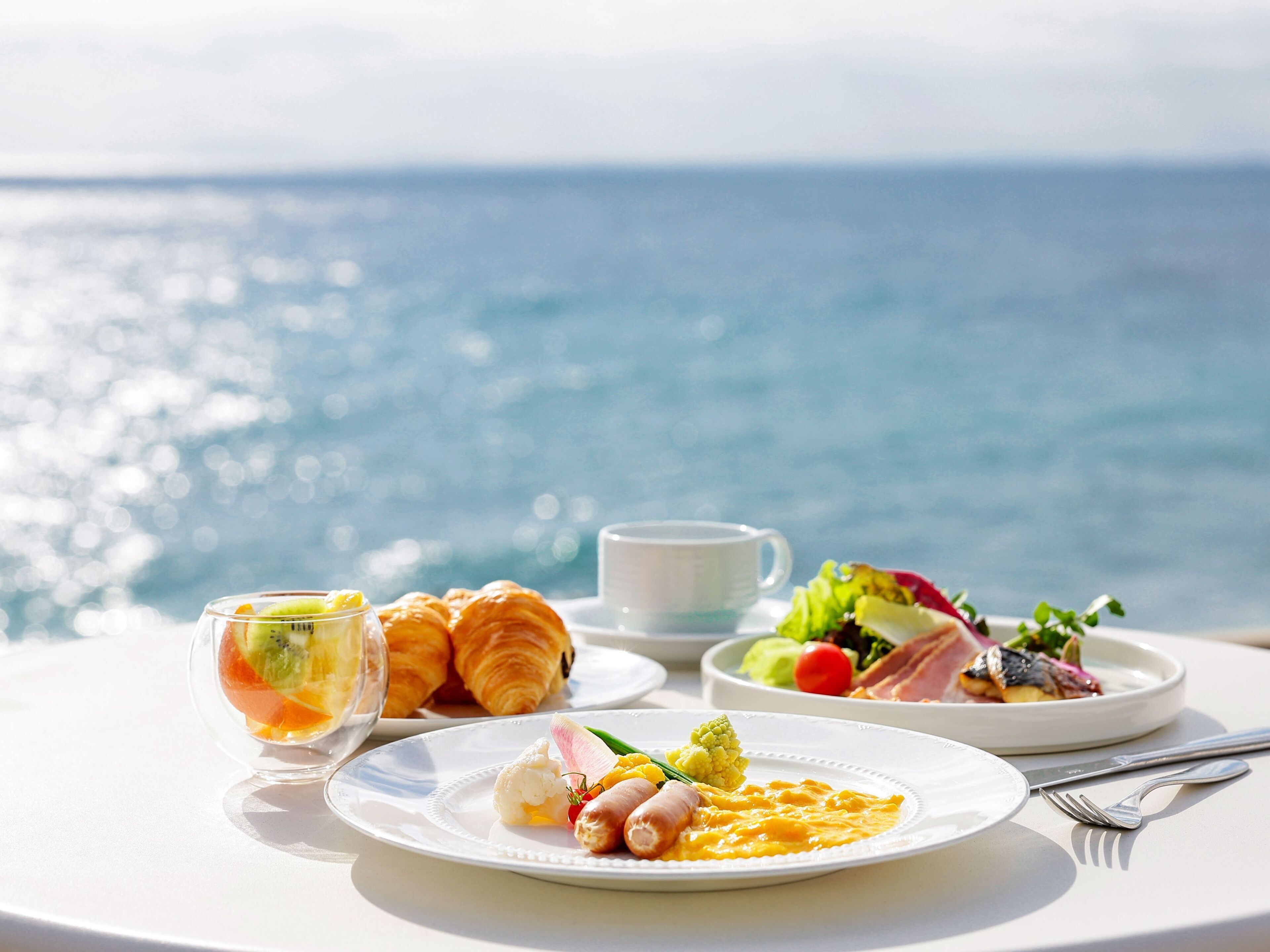A table with breakfast items including croissants, a glass of juice, and a plate with eggs and sausages, set against a scenic ocean view.