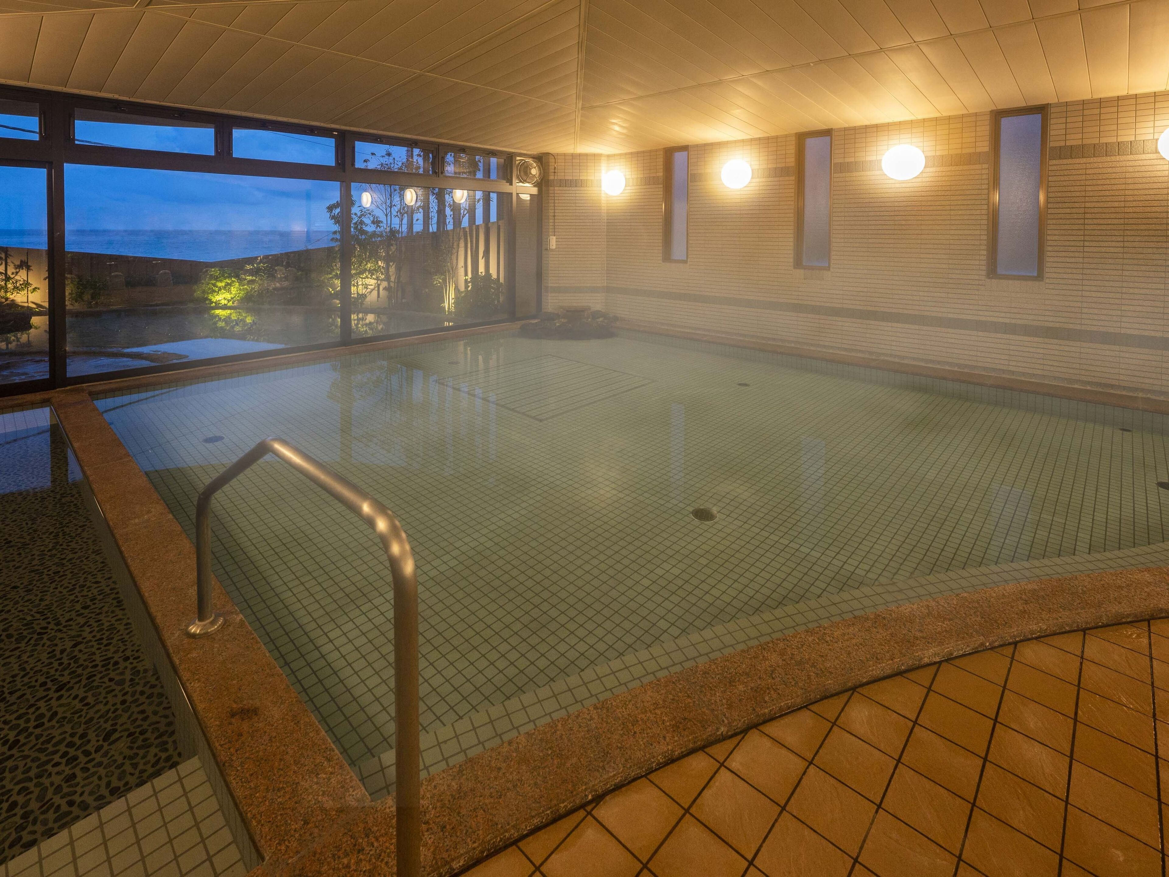 An indoor swimming pool with clear glass walls, a tiled floor, and a view of the outdoors at dusk.