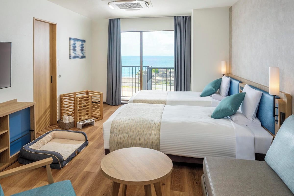 Habitación de hotel con dos camas, un televisor, un escritorio, una mesita pequeña y un balcón con vistas.